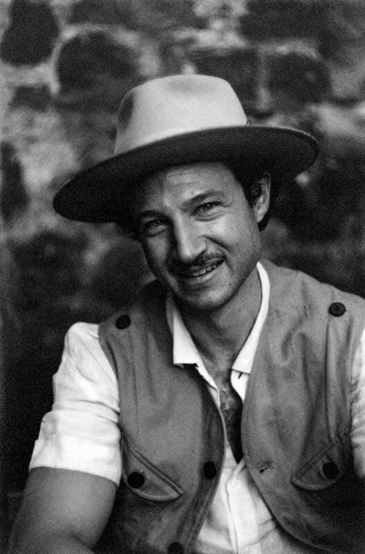 A man wearing a wide-brimmed hat, a light shirt, and a vest, smiles while looking at the camera. The background is blurred with a stone or brick wall pattern. The image is in black and white.