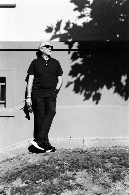 An older man wearing sunglasses, a dark shirt, and pants stands casually against a sunlit wall with shadows of leaves. He holds something in his hand and smiles, appearing relaxed and content.