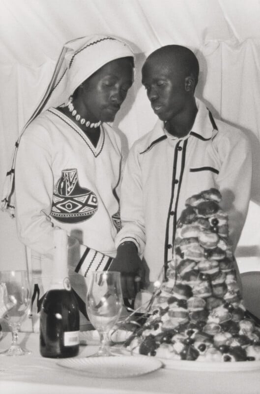 Two people dressed in patterned traditional and formal attire stand together, cutting a cake at a decorated table with a champagne bottle, glasses, and a tall stack of pastries.