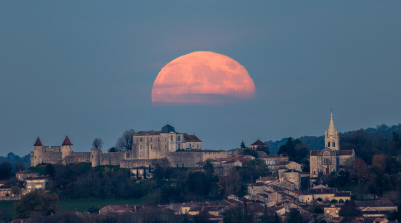 A large, orange full moon rises over a historic hilltop village with stone buildings, a church, and a castle, surrounded by trees at dusk.