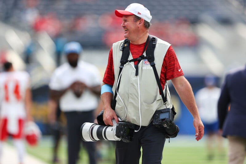 A photographer in a gray vest and red cap is smiling on a football field, carrying a camera with a long lens and another camera over his shoulder. Blurred players and spectators are visible in the background.