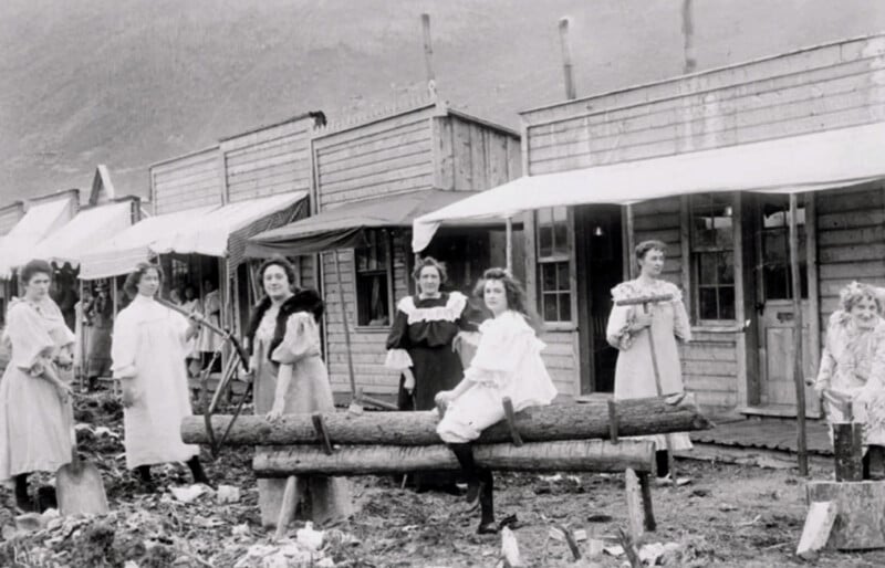Seven women in old-fashioned dresses pose outside wooden buildings in a rustic, muddy street. Two sit on a log in the foreground, while others stand nearby. The scene appears to be from a historical frontier town.