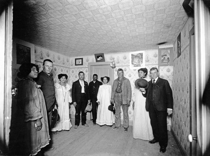 A group of nine people, men and women, stand in a small, wallpapered room with framed pictures on the walls. Some hold hats and wear formal or period clothing, posing for a black-and-white photograph.