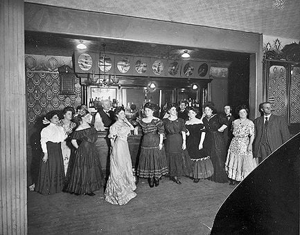 A vintage black-and-white photo shows a group of women in formal dresses and one man in a suit standing in front of a bar inside an ornately decorated room, likely from the early 20th century.