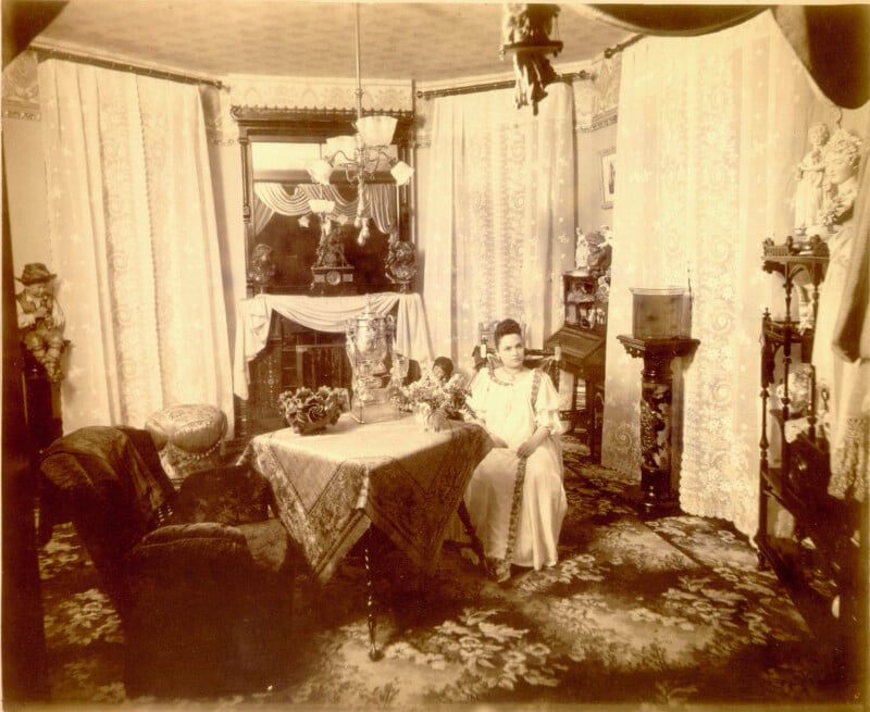 A sepia-toned photo of a Victorian-era living room with ornate furniture, lace curtains, floral carpet, and a woman in a long dress seated at a table decorated with flowers and ornaments.