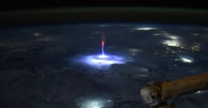 A view from space shows a bright blue and purple lightning sprite shooting upward above clouds over Earth, with city lights below and a spacecraft part visible in the foreground.