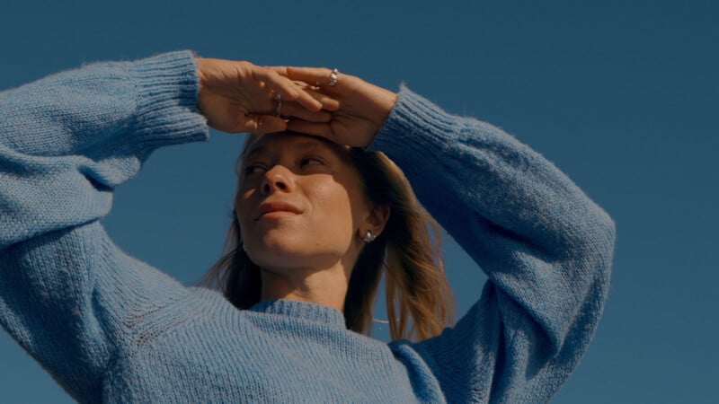 A woman in a blue sweater stands outdoors under a clear blue sky, gazing upward with her hands shielding her eyes from the sunlight.