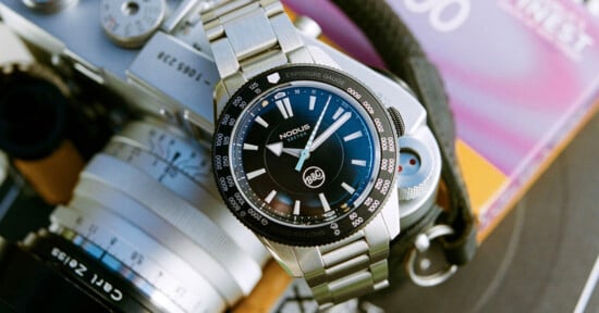 A stainless steel wristwatch with a black face and luminous markings is placed on a vintage camera. The watch has two sub-dials and a branded logo. The background includes a colorful, blurred book cover.