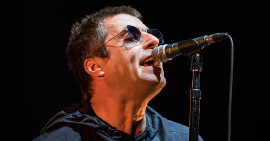 A man wearing sunglasses and a dark jacket sings into a microphone on stage against a black background.