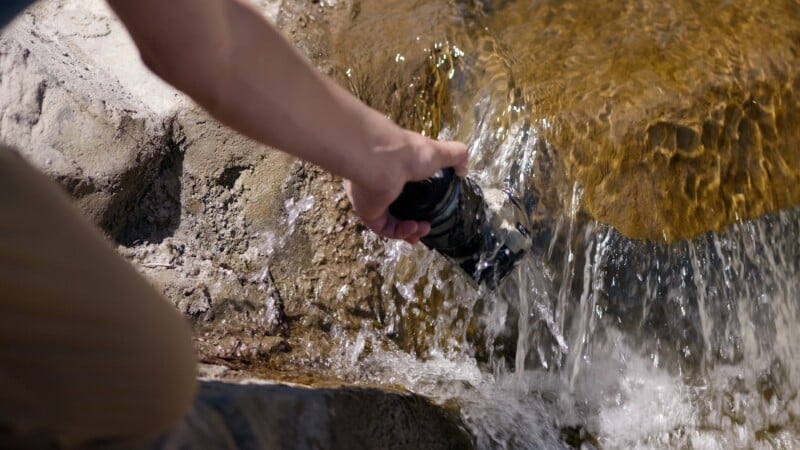 A person fills a black water bottle from a clear mountain stream, holding it under flowing water against a rocky surface.