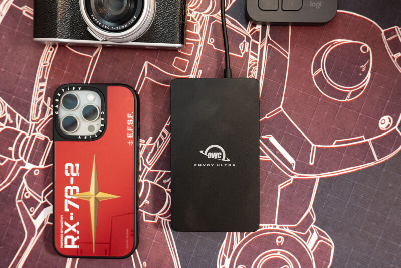 A smartphone with a red RX-78-2 Gundam-themed case, an external OWC Envoy Ultra drive, and a camera rest on a mat with white mechanical drawings.