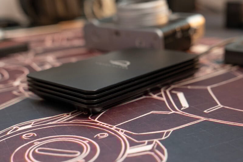 A close-up of a sleek black external hard drive with rounded edges resting on a surface featuring a robot illustration, with a blurred camera in the background.