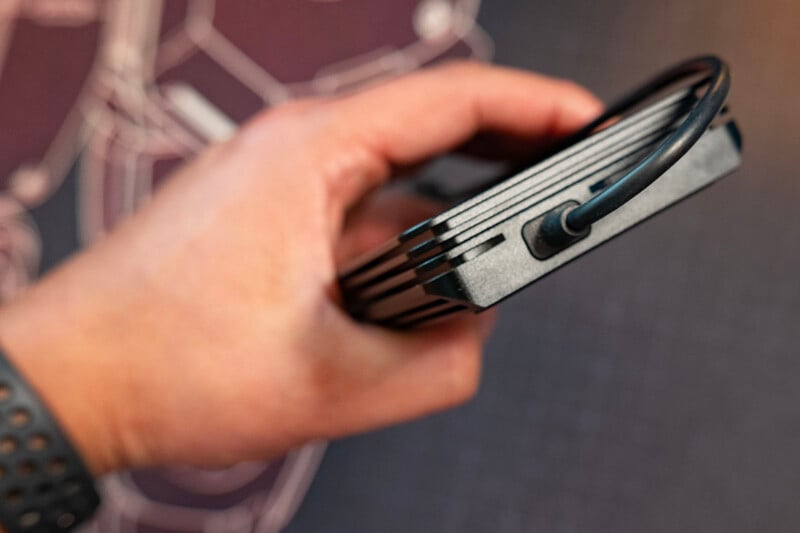 A hand holding a rectangular black electronic device with a cable connected to one end, against a blurred background.
