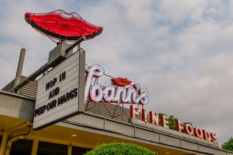 A retro diner sign with red lips and neon lights reads "Joann's Fine Foods." A marquee below says, "HOP IN AND PEEP OUR MARGS." The sky is partly cloudy above the building.
