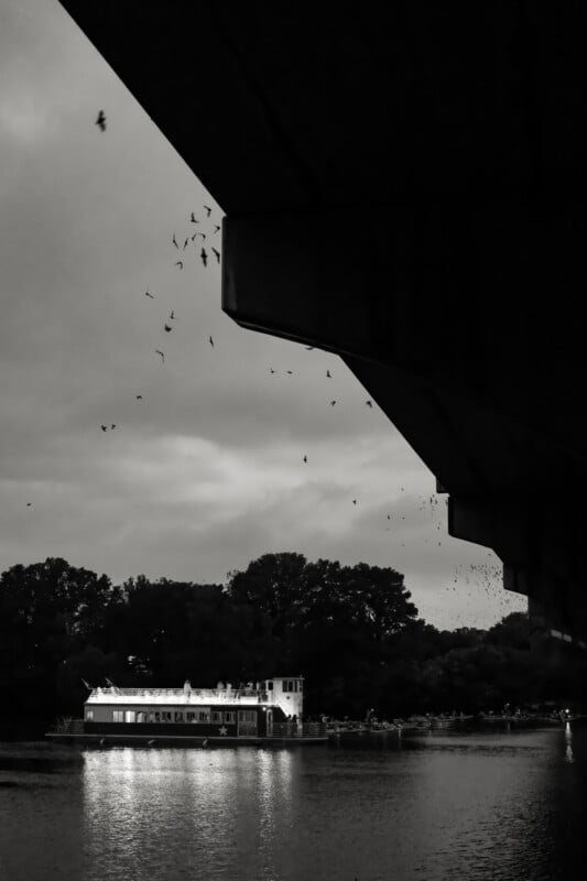 A brightly lit boat is moored on a river at dusk, with trees in the background. The dark silhouette of a bridge dominates the foreground, and birds are flying in the sky. The image is in black and white.