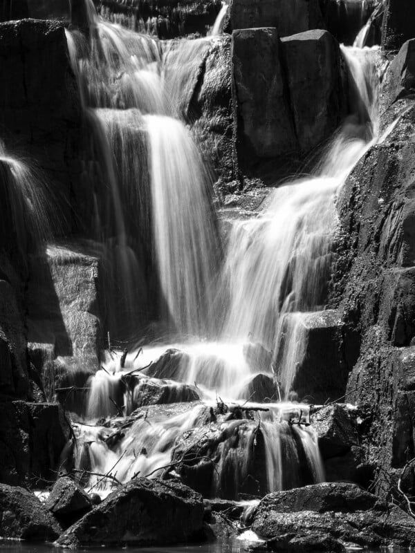 A black and white photo of a multi-tiered waterfall cascading over rocks, with water flowing smoothly and creating a soft, misty effect among the rugged, textured stones.