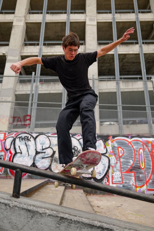 A skateboarder in black clothes performs a trick on a handrail in front of a building with graffiti-covered walls and a metal fence.