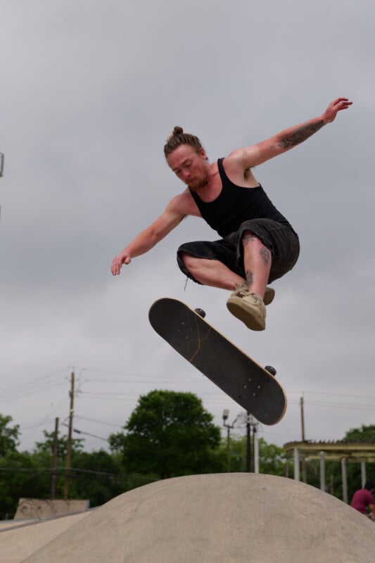 A skateboarder with a man bun and tattoos, wearing a black tank top and shorts, performs a mid-air trick over a concrete ramp at a skate park under a cloudy sky.