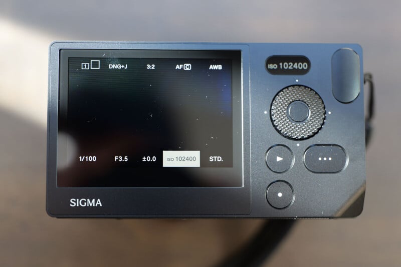 The image shows the back of a Sigma camera, focusing on the screen that displays image settings, including DNG+J format, ISO 102400, shutter speed 1/100, aperture f/3.5, and autofocus in use. Several buttons are visible around the screen.