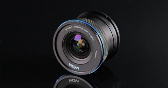 A Laowa camera lens with a wide-angle view sits on a reflective black surface, showing the front glass element and engraved details for focal length and aperture around the rim.