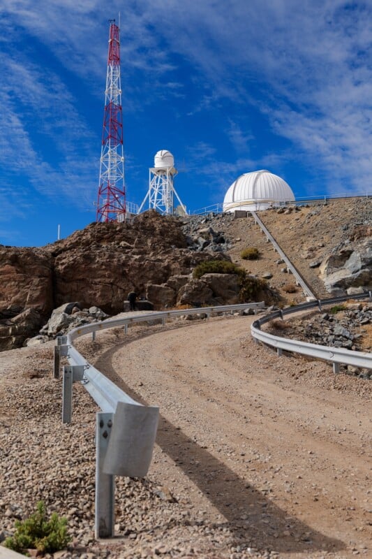 A winding dirt road with metal guardrails leads up to a white observatory dome and a tall red-and-white communication tower set on a rocky hillside under a blue sky with scattered clouds.