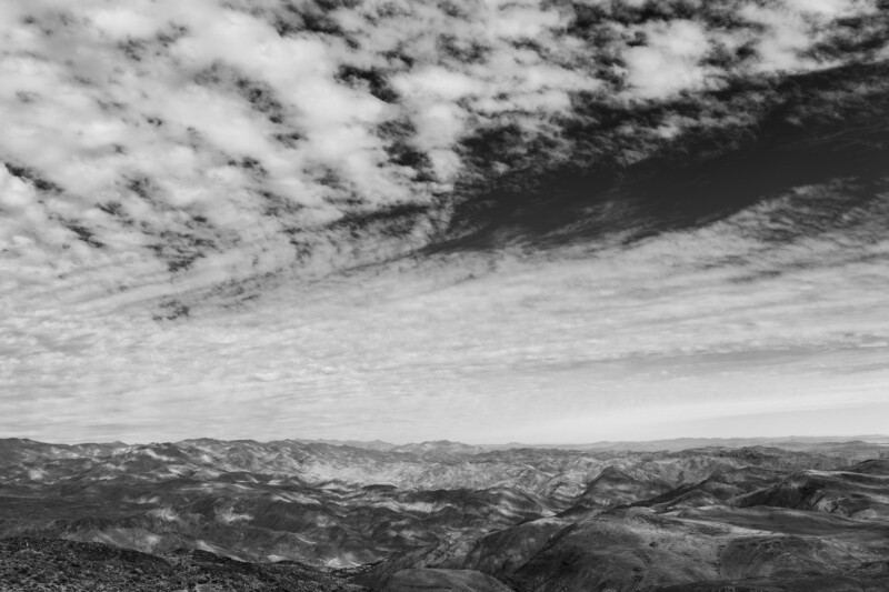 Black and white photo of a dramatic sky filled with streaky clouds above a vast, rugged mountain landscape, creating a sense of depth and contrast.
