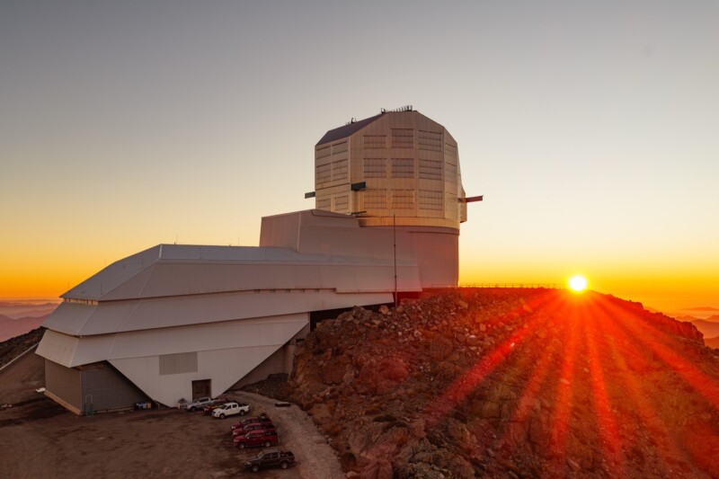 A large observatory sits atop a rocky mountain at sunset. The sun is low on the horizon, casting a warm glow and long rays of light. Several parked cars are visible near the building’s entrance.