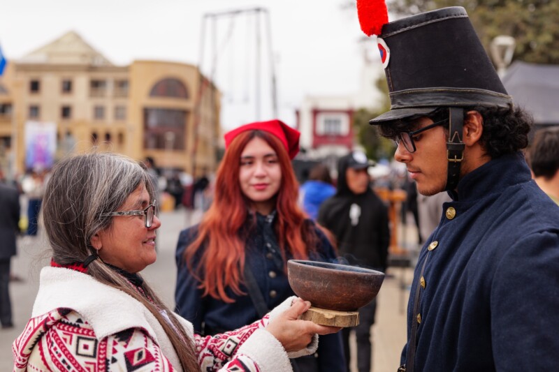 A woman in a patterned sweater offers a wooden bowl to a young man in a historical military uniform and tall black hat, while another young woman with long hair and a red hat watches in the background at an outdoor event.