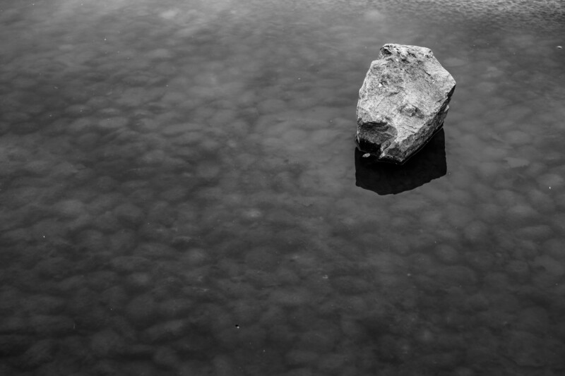 A single large rock rests in shallow water, its reflection faintly visible. The surface of the water is calm and slightly cloudy, creating a moody, minimalist scene in black and white.
