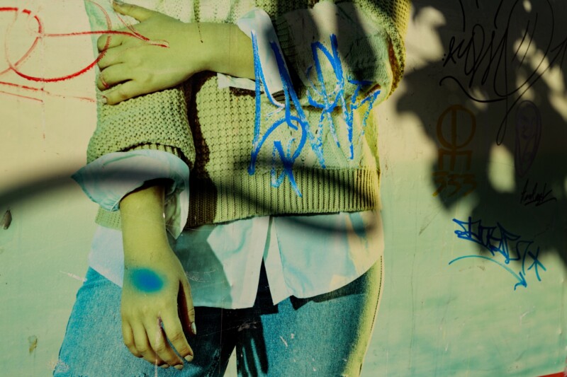 A person in a sweater and jeans stands against a wall with colorful graffiti, their arms crossed. Shadows and sunlight create dramatic patterns across their body and the wall.
