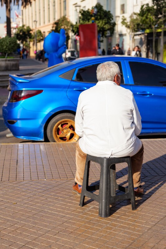 An older man with gray hair, wearing a white jacket and beige pants, sits on a black plastic stool facing away from the camera. A blue car and a blue statue are visible on the street in front of him.