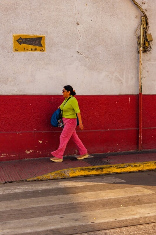 A woman in a bright green shirt and pink pants walks along a sidewalk beside a red and white wall, carrying a blue bag. A faded street sign is visible above her on the wall.