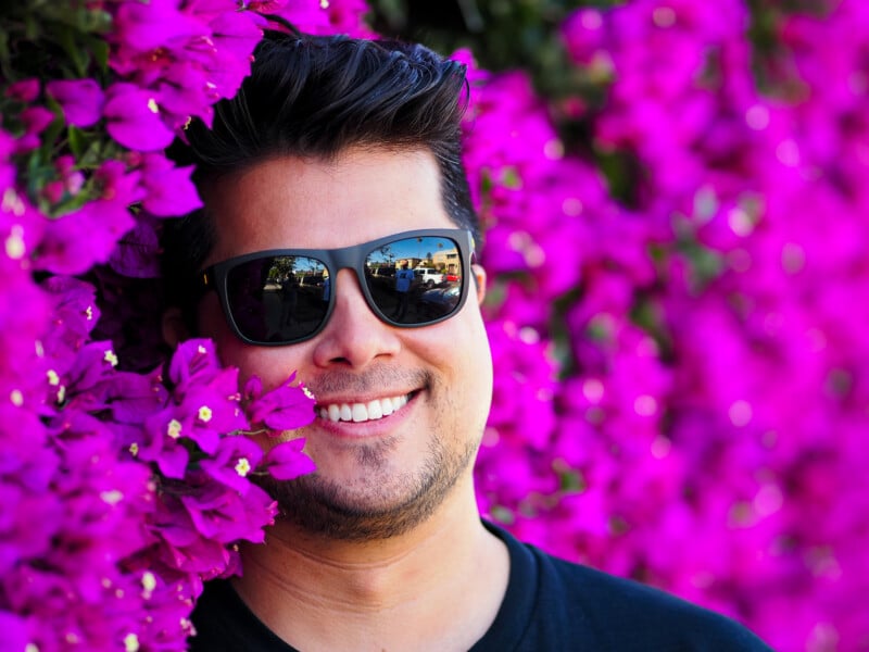 A smiling man wearing sunglasses stands close to vibrant pink bougainvillea flowers, with the blossoms surrounding his face. The background is filled with the same bright flowers.