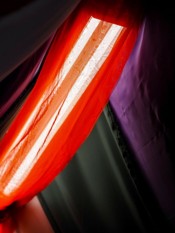 A bright beam of sunlight shines through sheer red and orange fabric draped next to purple and dark gray fabric, creating a colorful, layered effect.