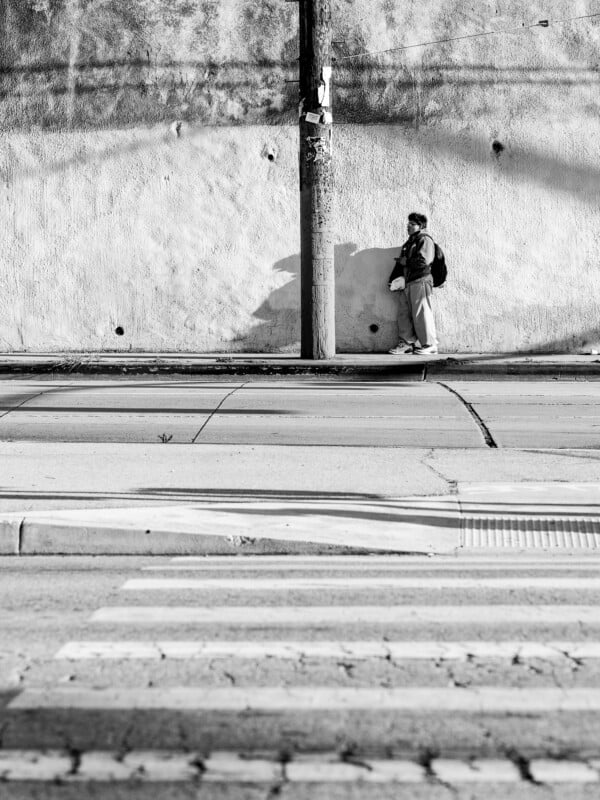 Black and white photo of a person standing alone against a textured wall, beside a pole, across an empty street with a crosswalk in the foreground.