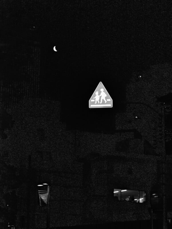 A triangular pedestrian crossing sign glows at night above a dark street, with the crescent moon visible in the sky. Some building outlines and streetlights can be seen in the shadows.