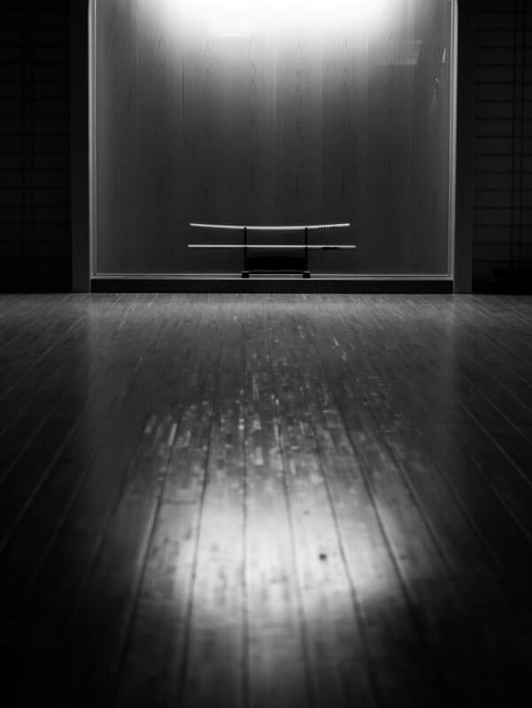 A black-and-white photo of three katana swords displayed on a stand against a softly lit wooden wall, with light reflecting off a polished wooden floor in the foreground.