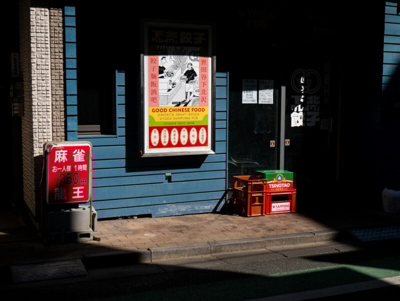 A sunlit street corner with a blue wooden building, a poster advertising "Good Chinese Food," a red crate with Tsingtao beer bottles, and a red sign displaying prices in Japanese. Shadows partially cover the entrance.