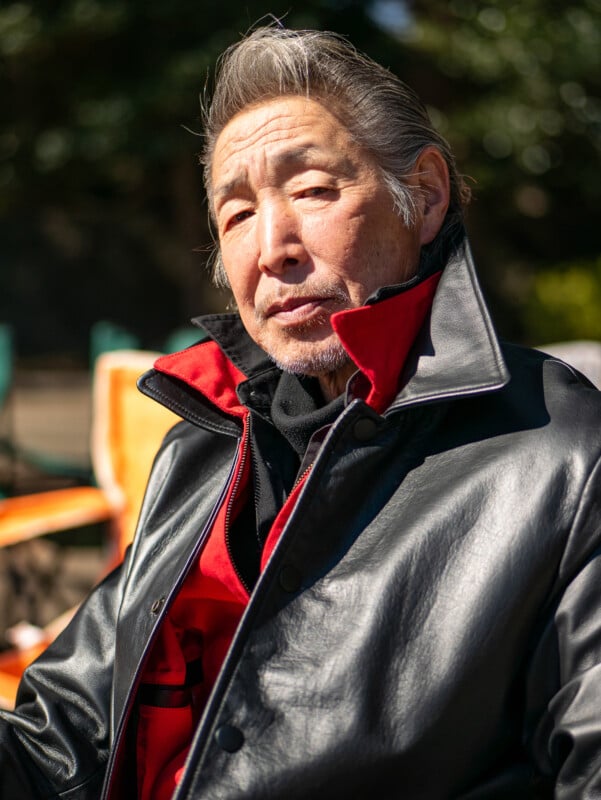 An older man with gray hair and a goatee wears a black leather jacket with a red inner lining, sitting outdoors on a sunny day, looking thoughtfully at the camera.
