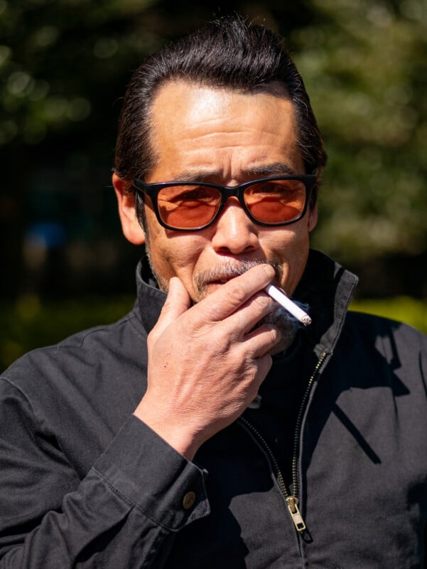 A man wearing amber sunglasses and a black jacket holds a cigarette near his mouth while standing outdoors in sunlight.
