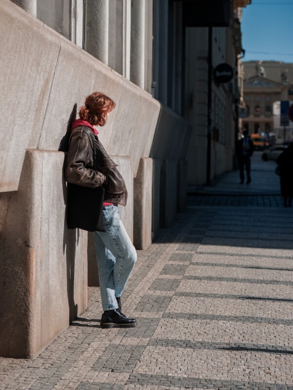 A person with short hair, wearing a leather jacket, jeans, and black shoes, leans against a concrete wall on a sunlit city sidewalk, casting a shadow. Other people are seen walking in the background.
