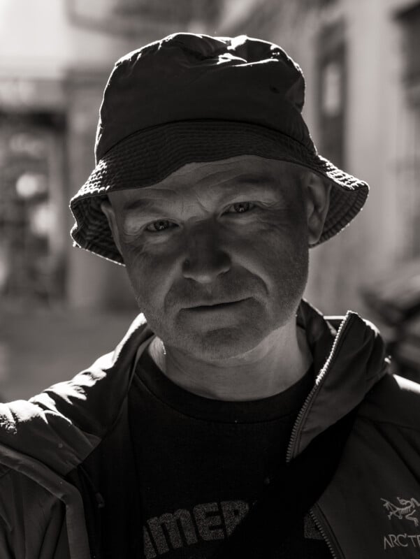 A man in a bucket hat and jacket stands outdoors, lit from the side by sunlight. His expression is neutral, and the image is in black and white with a blurred urban background.