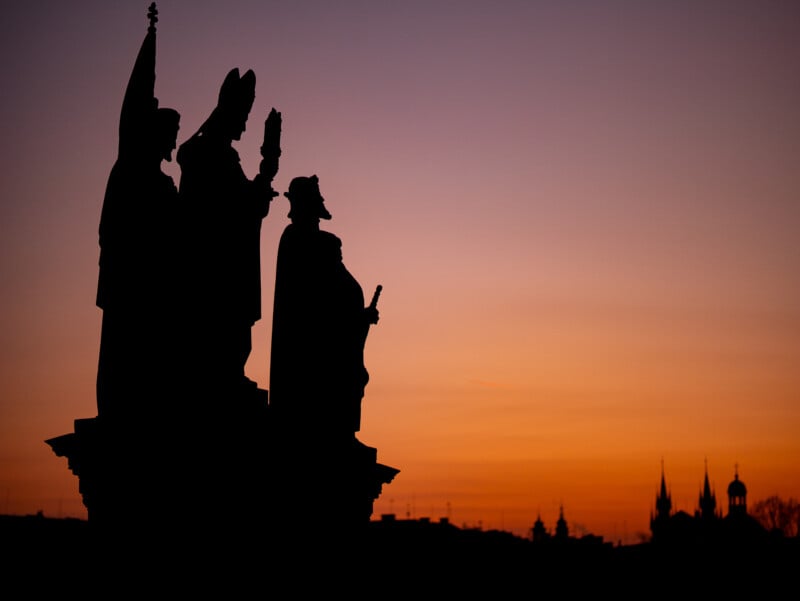 Silhouettes of three statues with raised arms stand against a vibrant orange and purple sunset sky, with distant building spires visible on the horizon.