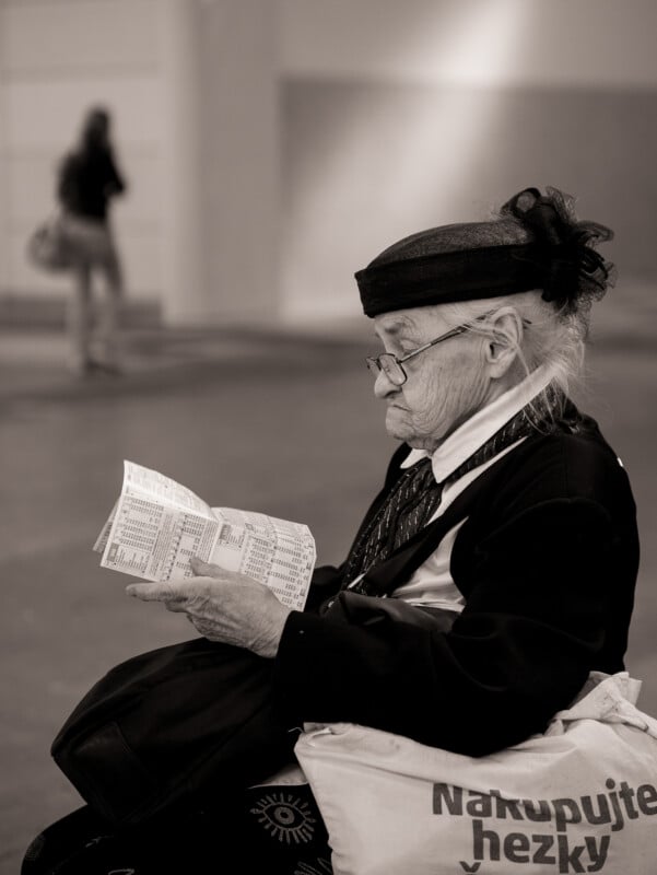 An elderly woman wearing glasses and a hat sits on a bench, reading a paper. She has a large bag on her lap and appears focused. The background is out of focus, with a person walking away. The photo is in black and white.