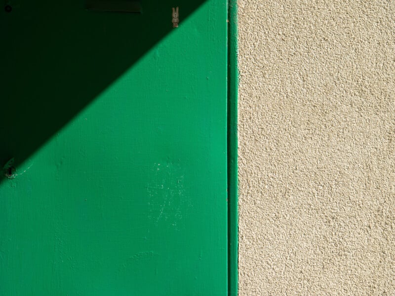 A green door partially in shadow stands next to a textured beige wall. The shadow creates a diagonal line across the door, dividing it from the sunlit wall.