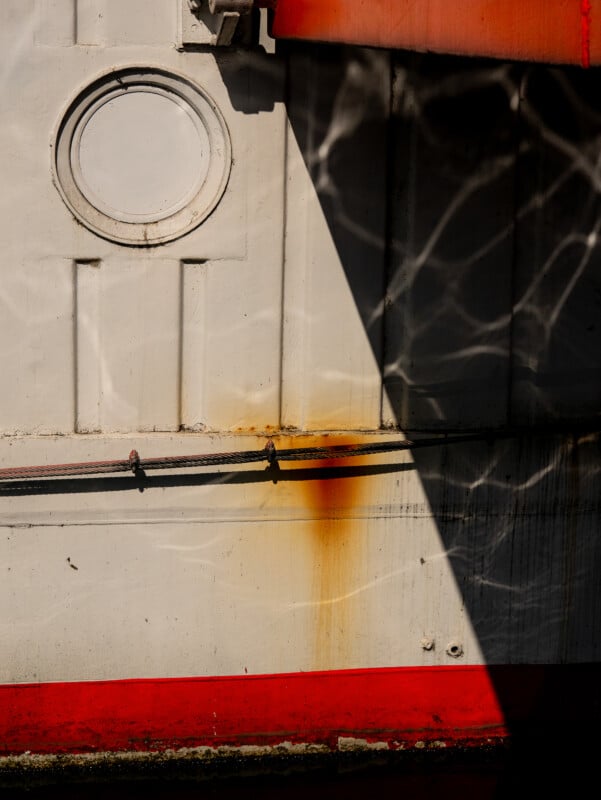 A close-up of a white metal boat wall with a round porthole, red stripe at the bottom, rust stain beneath the porthole, and dappled sunlight casting angular shadows and reflections across the surface.