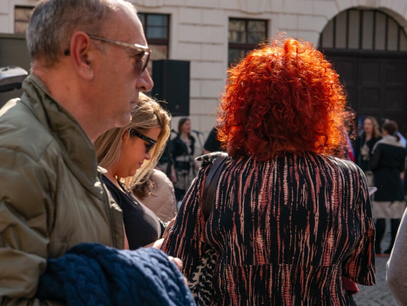 A man in sunglasses stands in profile, while a woman with bright curly red hair and others gather outdoors in sunlight near a building.