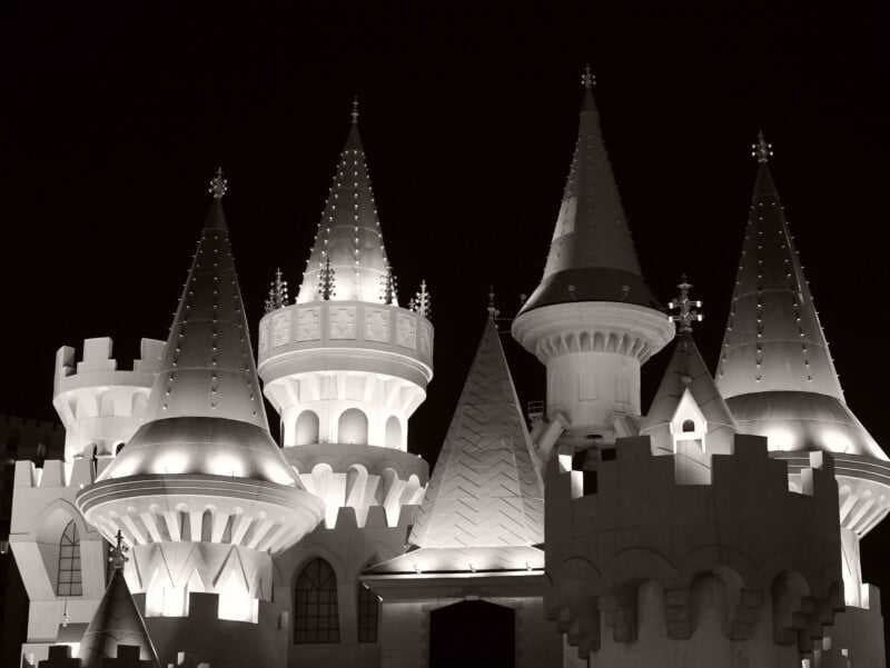 A black and white photo shows a whimsical castle with multiple pointed, turreted towers illuminated against the night sky. The architecture features fantasy-inspired details and decorative crenellations.