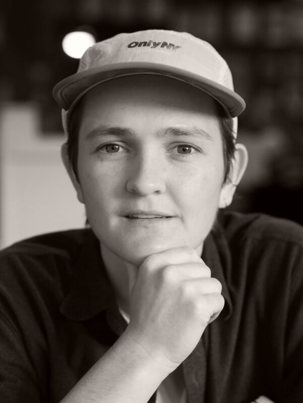 A person wearing a light-colored cap and dark shirt sits indoors, looking directly at the camera with their chin resting on their hand. The image is in black and white.