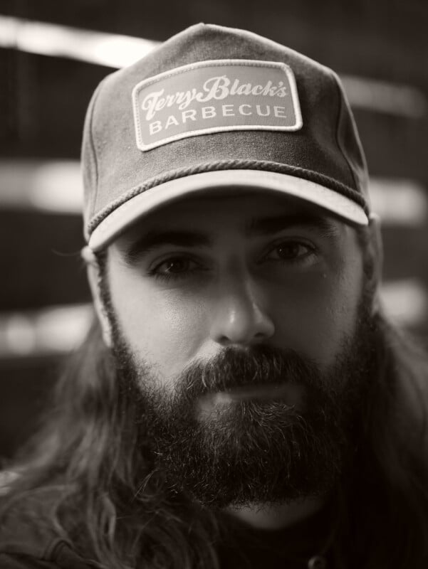 A bearded man with long hair wearing a "Terry Black's Barbecue" hat looks at the camera in a black-and-white portrait. The background is blurred.